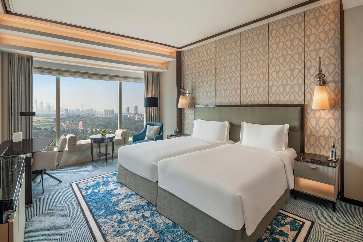 A hotel room with two beds, a desk, a chair, and a view of the city.