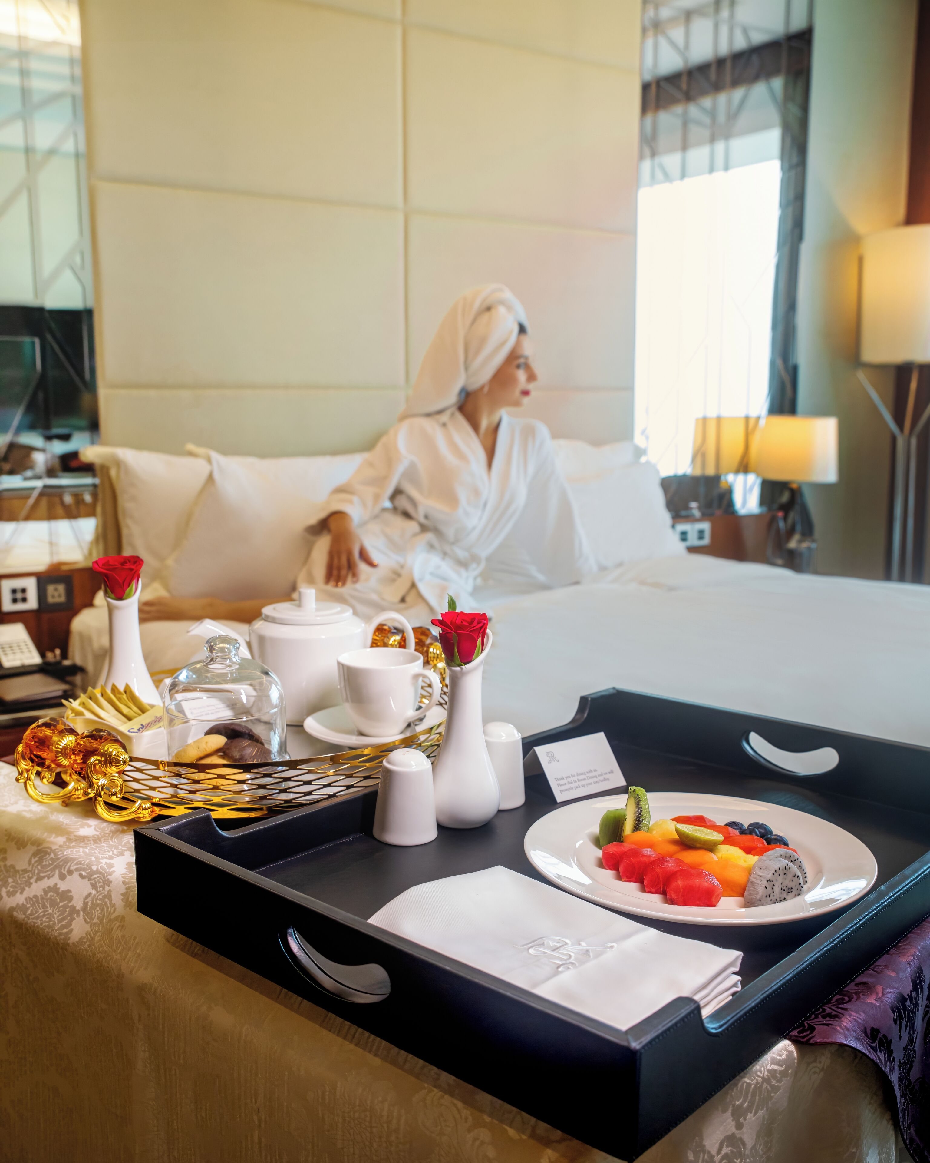 A hotel room with a bed, a tray with breakfast items, and a vase with a red rose.
