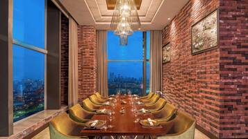 Comedor con una larga mesa dispuesta para seis comensales, paredes de ladrillo y vistas a la ciudad a través de amplias ventanas.