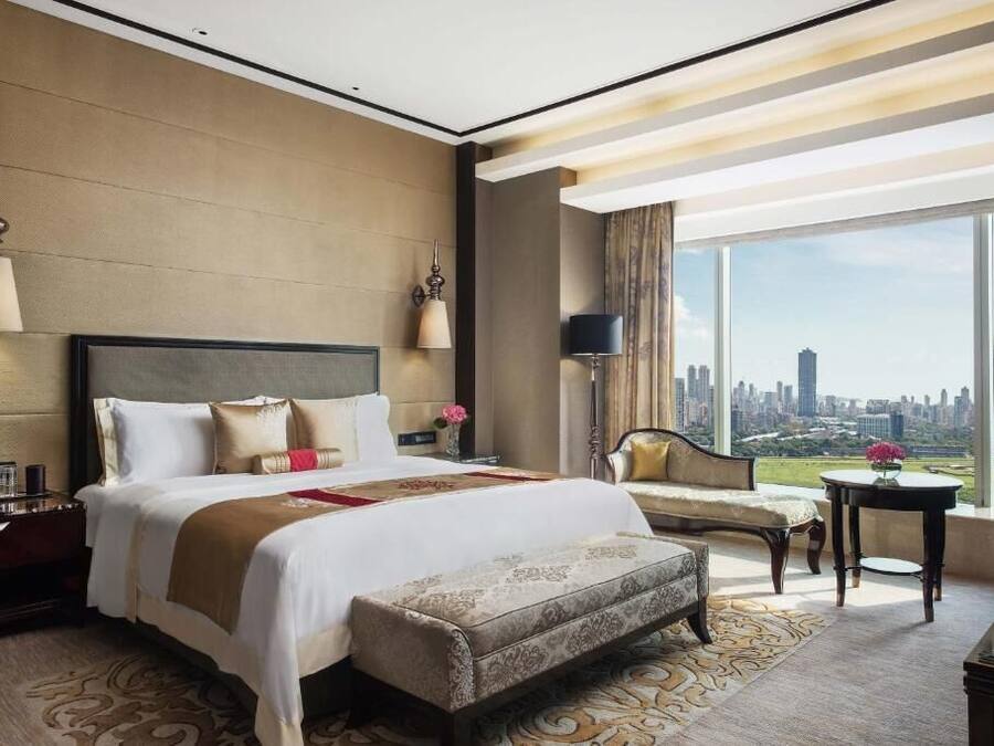 A hotel room with a large bed, a sofa, a small table, and a view of a cityscape.
