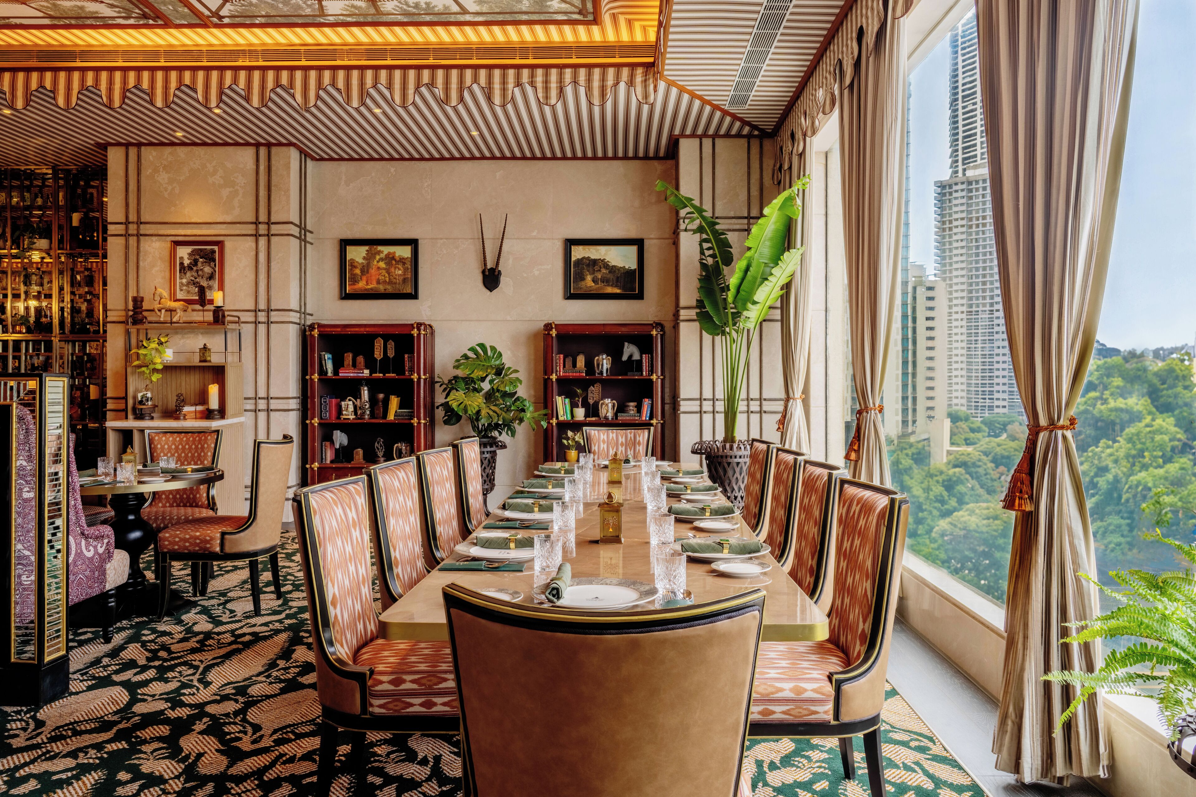 A dining room with a long table set for a meal, surrounded by chairs, with bookshelves and a large window offering a city view.