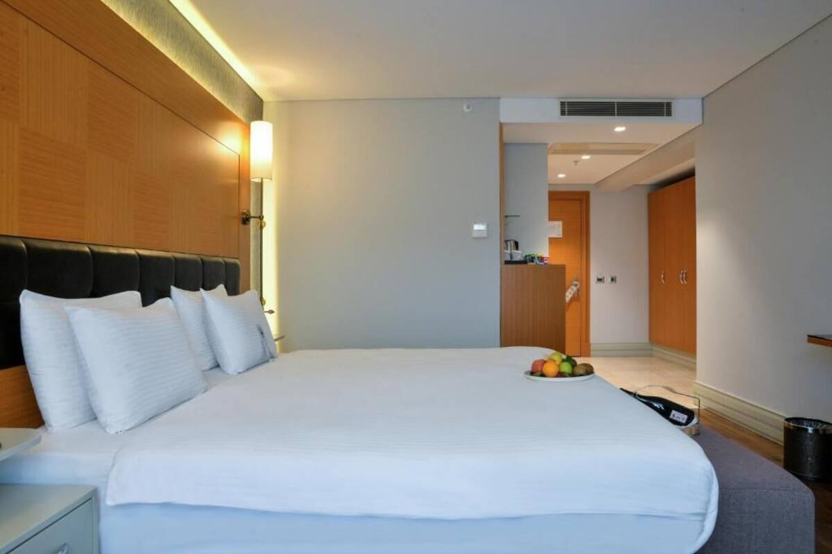 A hotel room with a large bed, a wooden headboard, a bedside table, and a fruit bowl on the bed.
