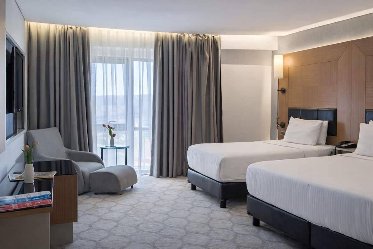 A hotel room with two beds, a chair, a small table with a vase, and a view of the city through large windows.