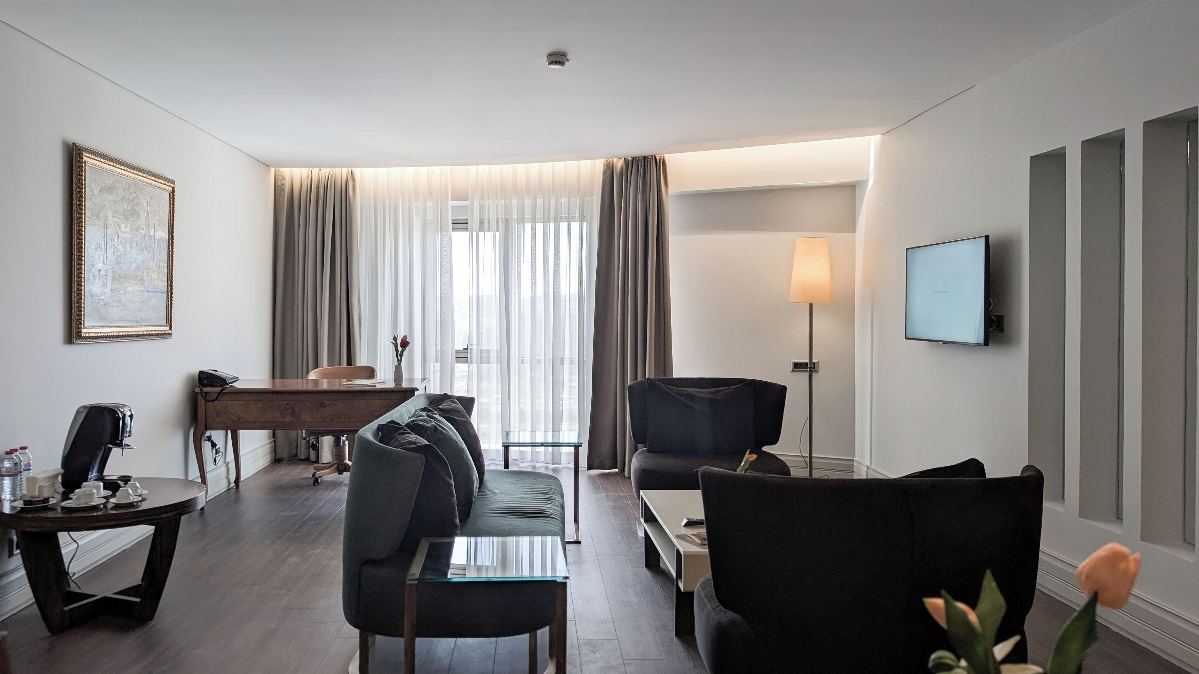 A modern hotel room with a living area, a dining table, a sofa, a coffee table, a TV, and a lamp.