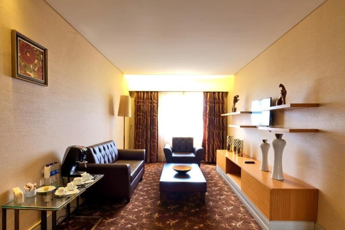 A hotel room with a sofa, armchair, coffee table, and a small desk with a lamp and decorative items.