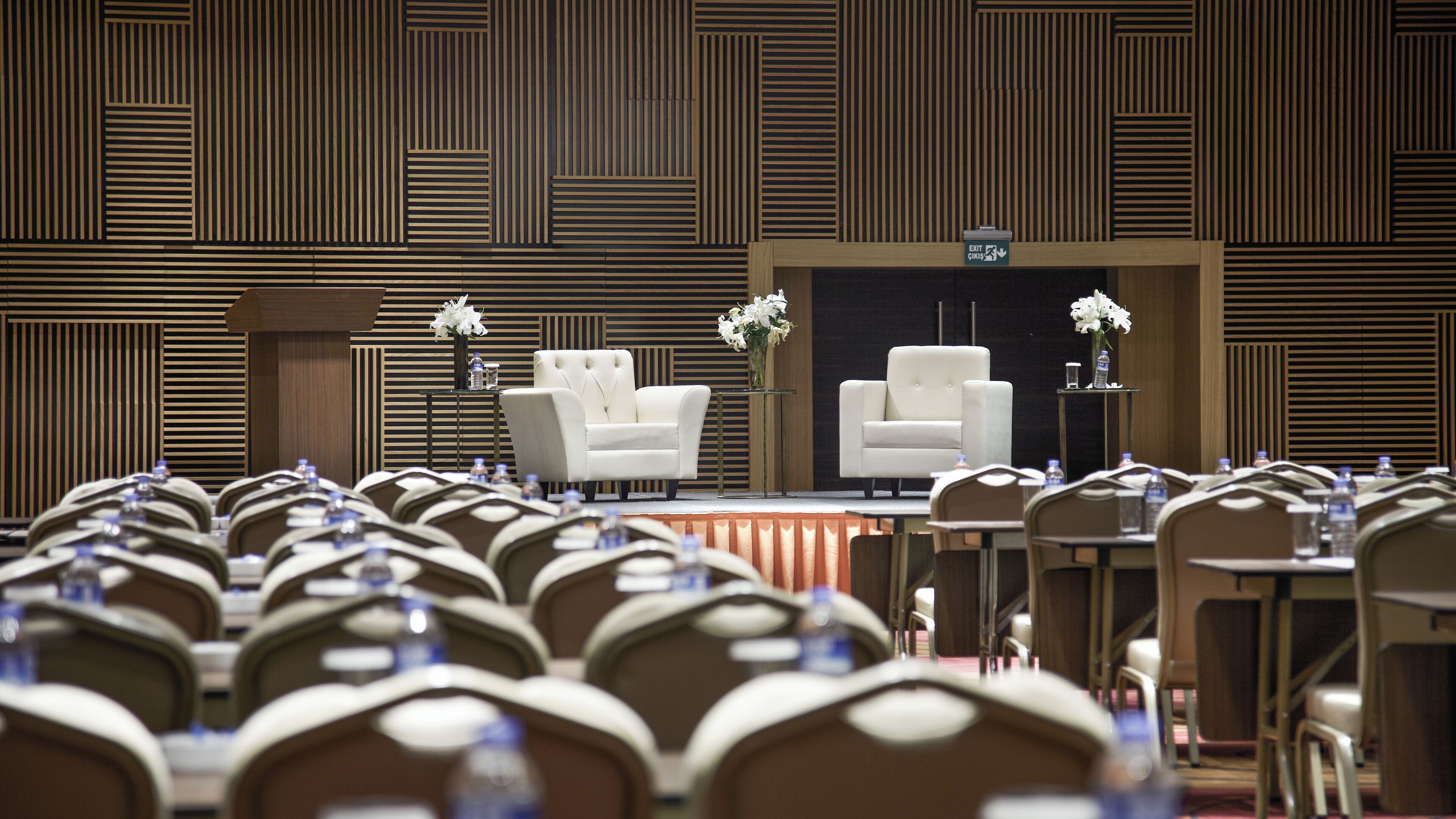 A conference room with rows of chairs, a stage with a white sofa and two vases of white flowers, and a wooden panel wall with vertical lines.