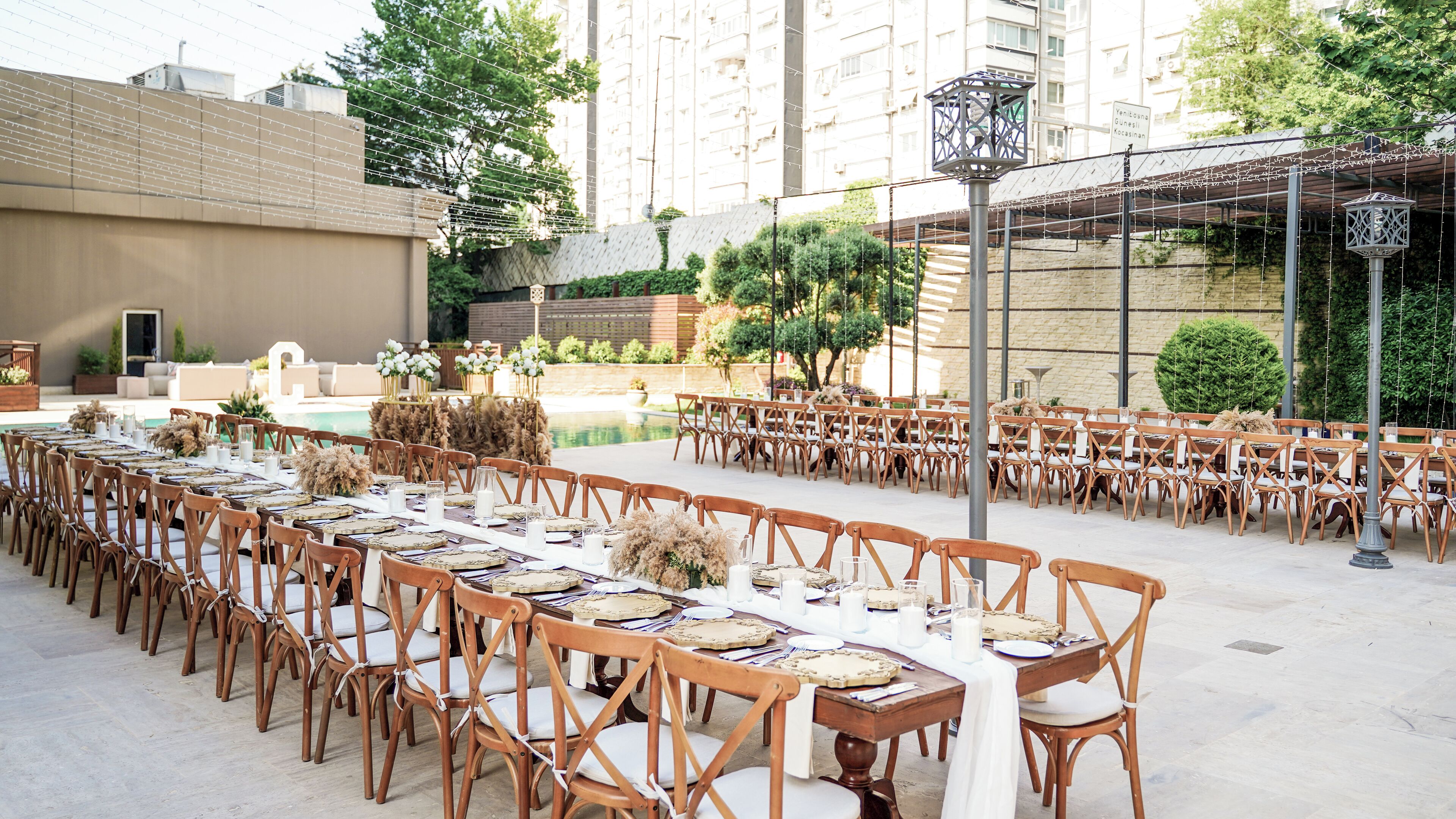 An outdoor dining area with long tables and wooden chairs, surrounded by greenery and a pool.