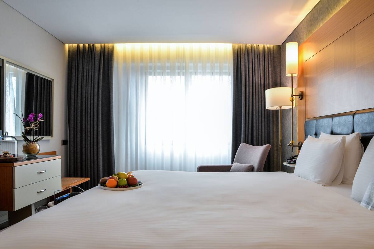 A hotel room with a large bed, a bedside table with a lamp, a chair, and a view of the outdoors through the window.