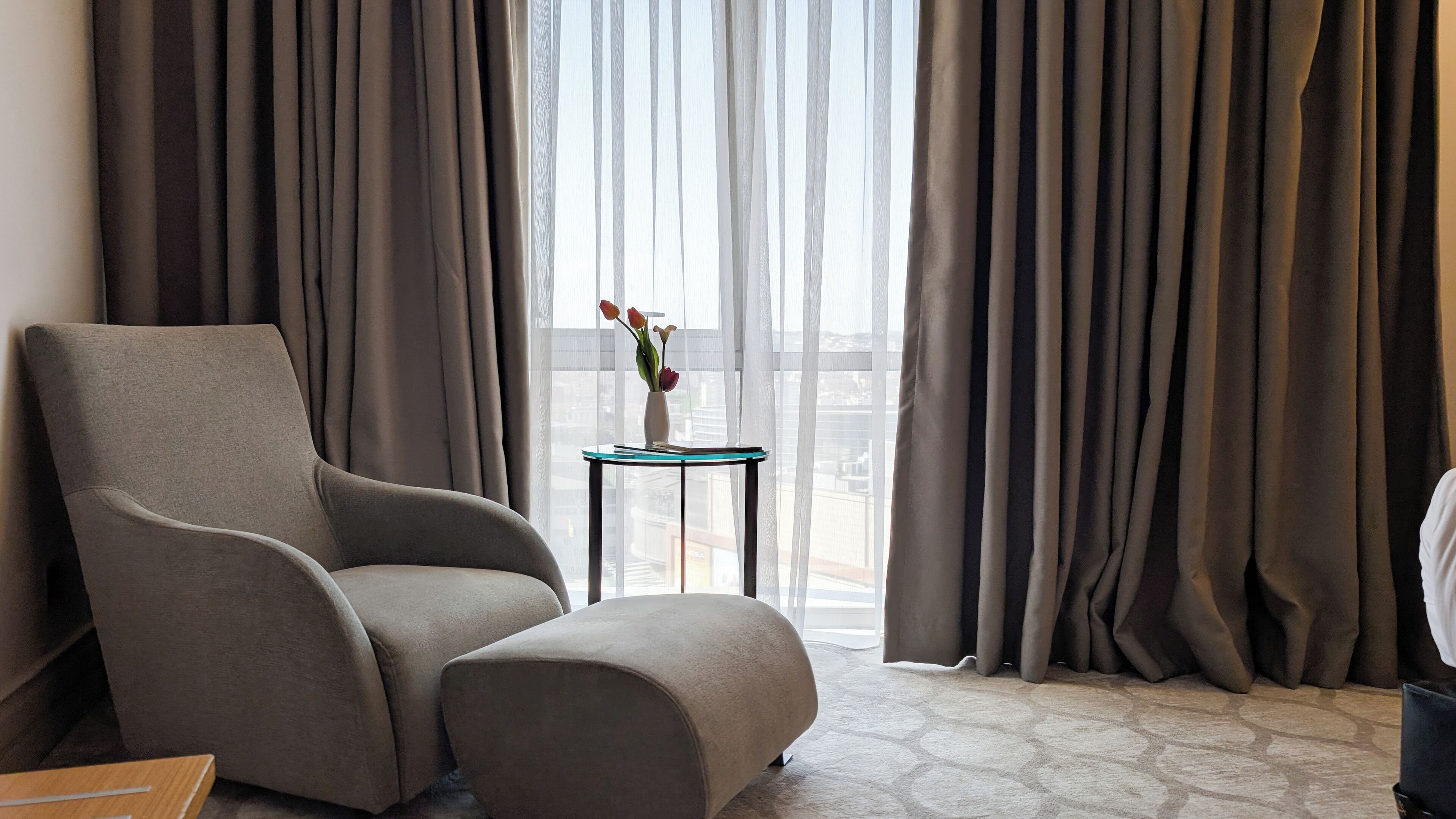 A modern hotel room with a grey armchair, a small side table with a vase of flowers, and a view of the city through sheer curtains.