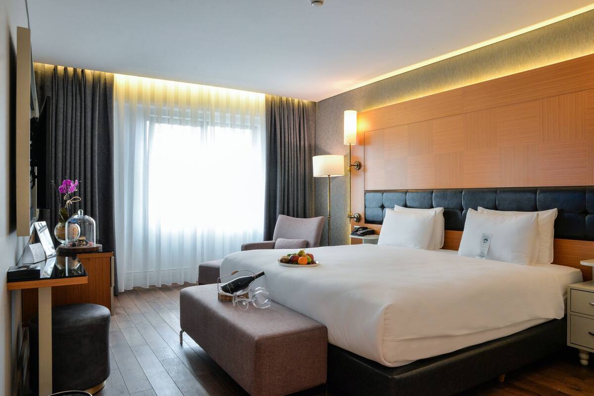 A hotel room with a large bed, a desk with a lamp, a chair, and a small table with a bottle and glasses.