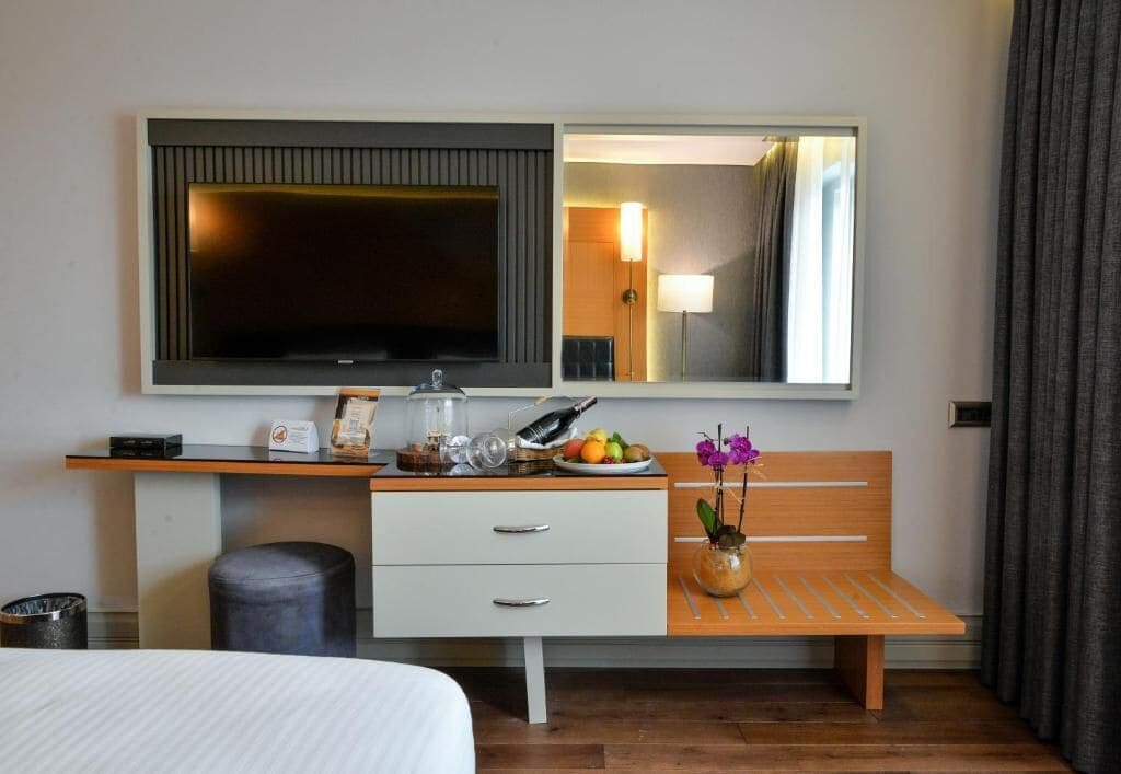 A hotel room with a bed, a desk with a flat-screen TV, a mirror, a wooden bench, and a small table with a vase of flowers.