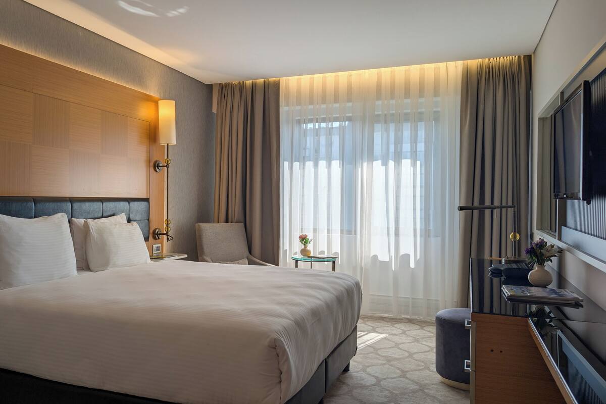 A hotel room with a large bed, a bedside table, a chair, a TV, and a window with curtains.