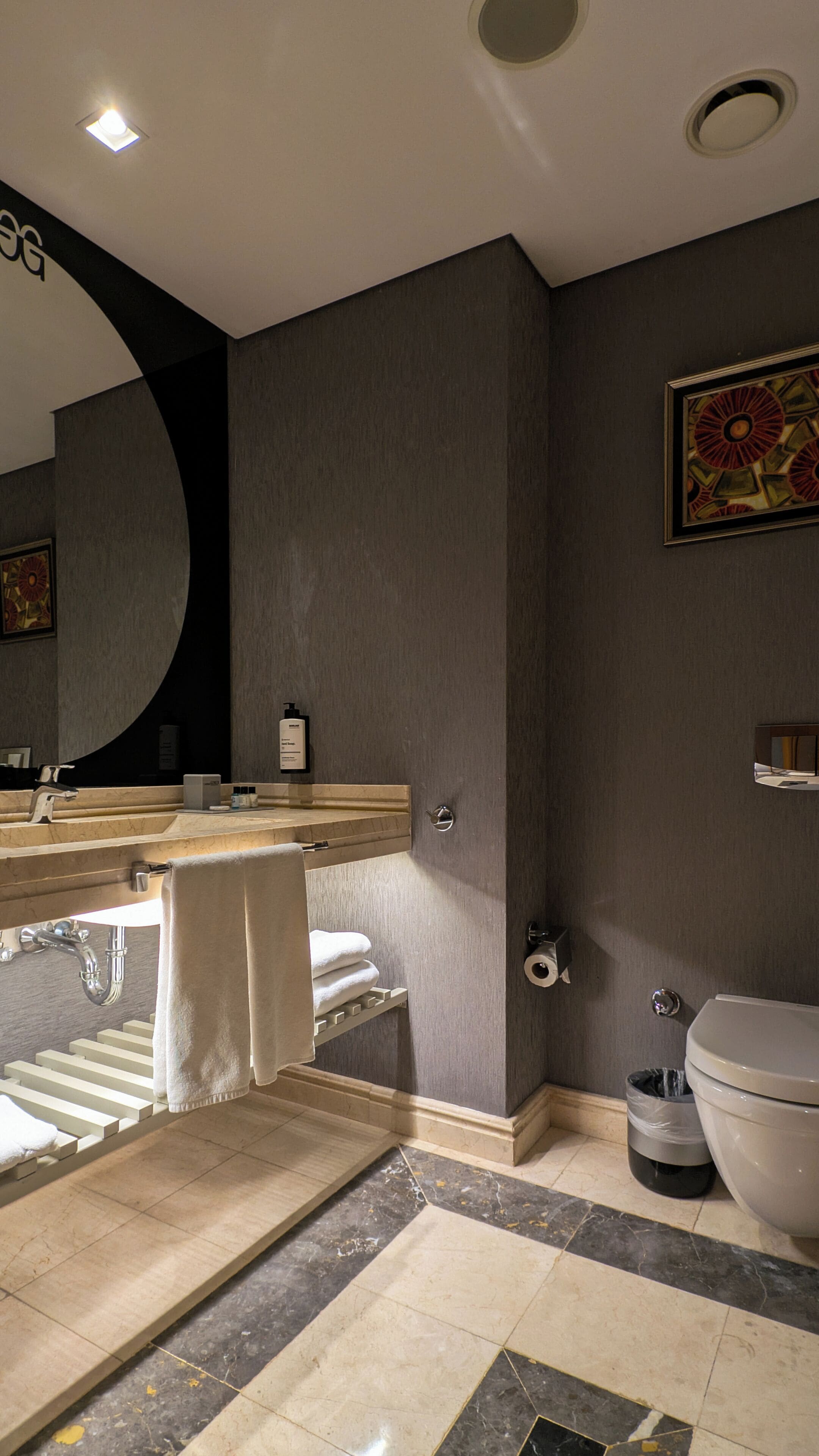 A modern bathroom with a large mirror, a sink with a towel rack, a toilet, and a trash bin.