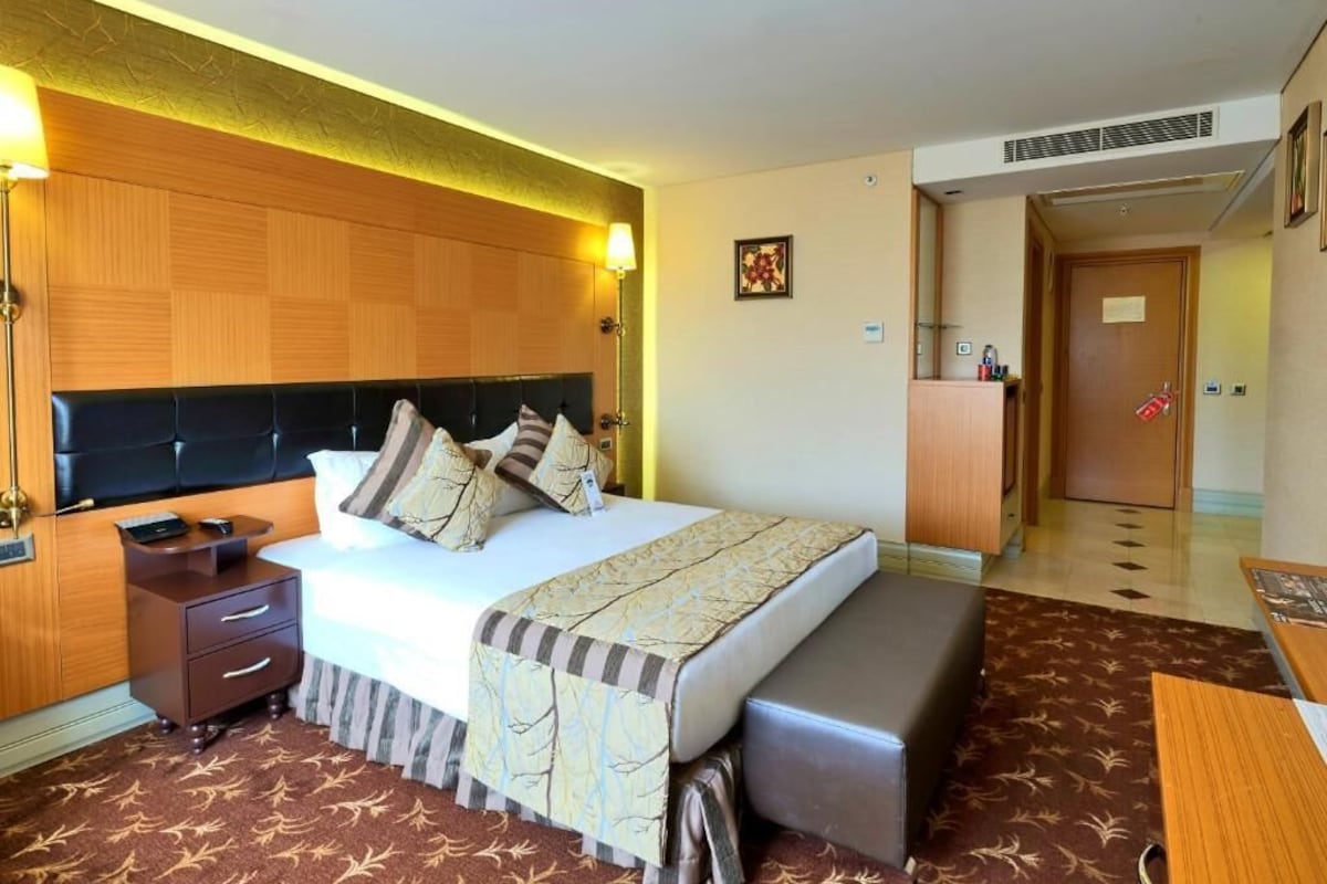 A hotel room with a bed, bedside table, lamp, and a small desk with a chair.