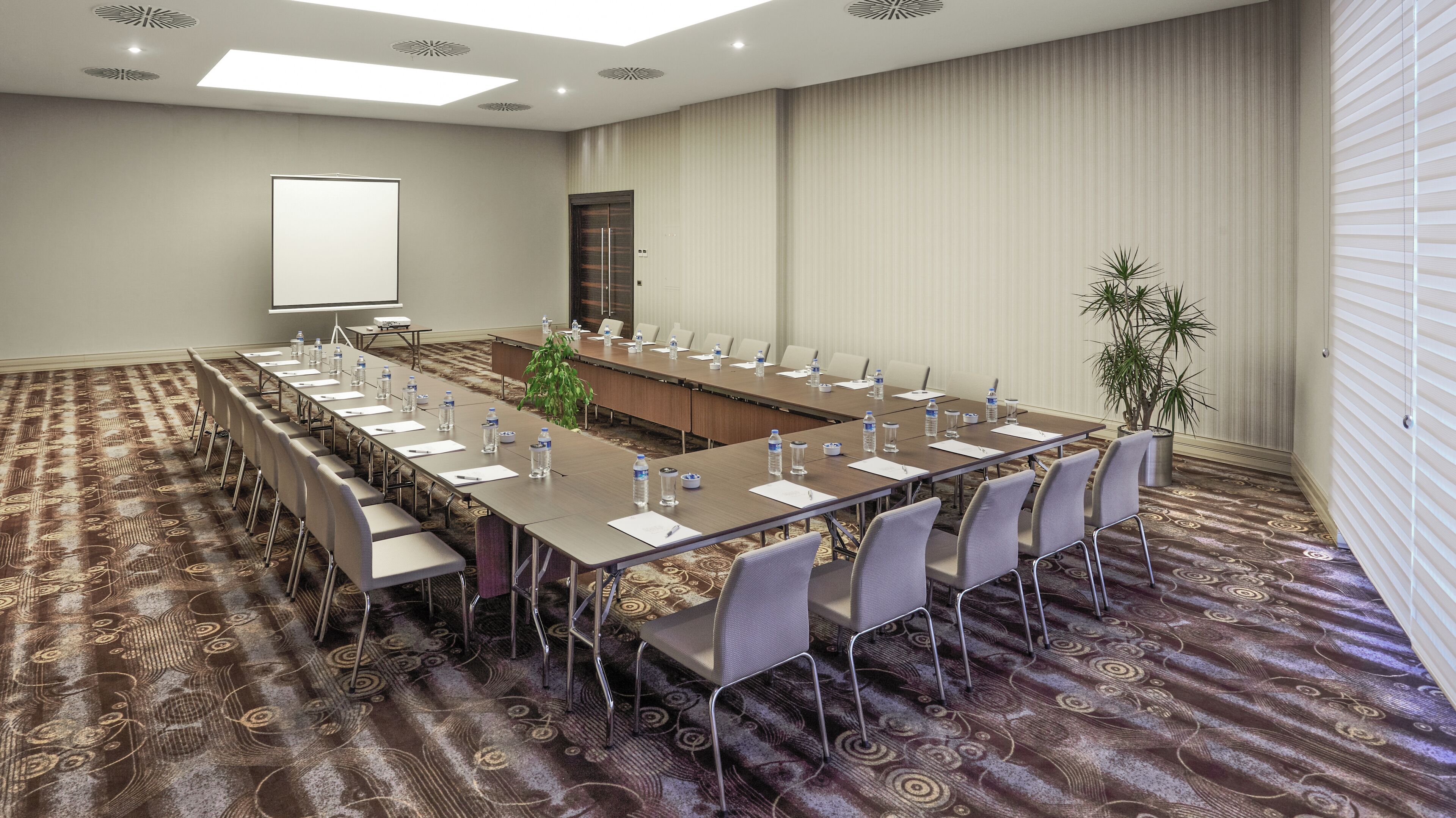 A conference room with a long table, chairs, a projector screen, and a plant.