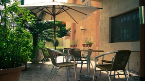 Un espace extérieur aménagé en terrasse, avec des chaises et une table en métal, entouré de plantes en pot et d’un grand parasol.