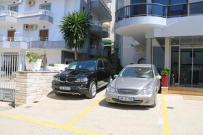 A parking area with two cars, a building with balconies, and a palm tree. - Brilant (Sarandë)