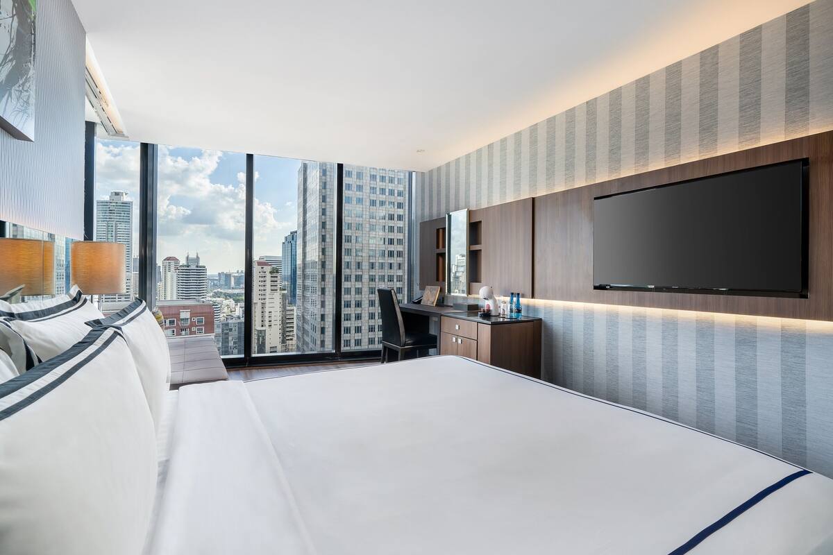 A modern hotel room with a large bed, a desk, and a TV. The room has large windows offering a city view.