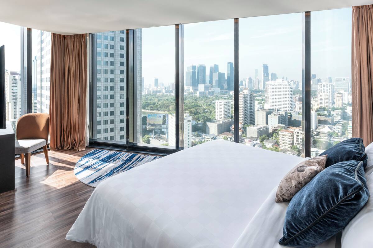 A modern hotel room with a large bed, a chair, a view of the cityscape through floor-to-ceiling windows, and a city skyline visible outside.