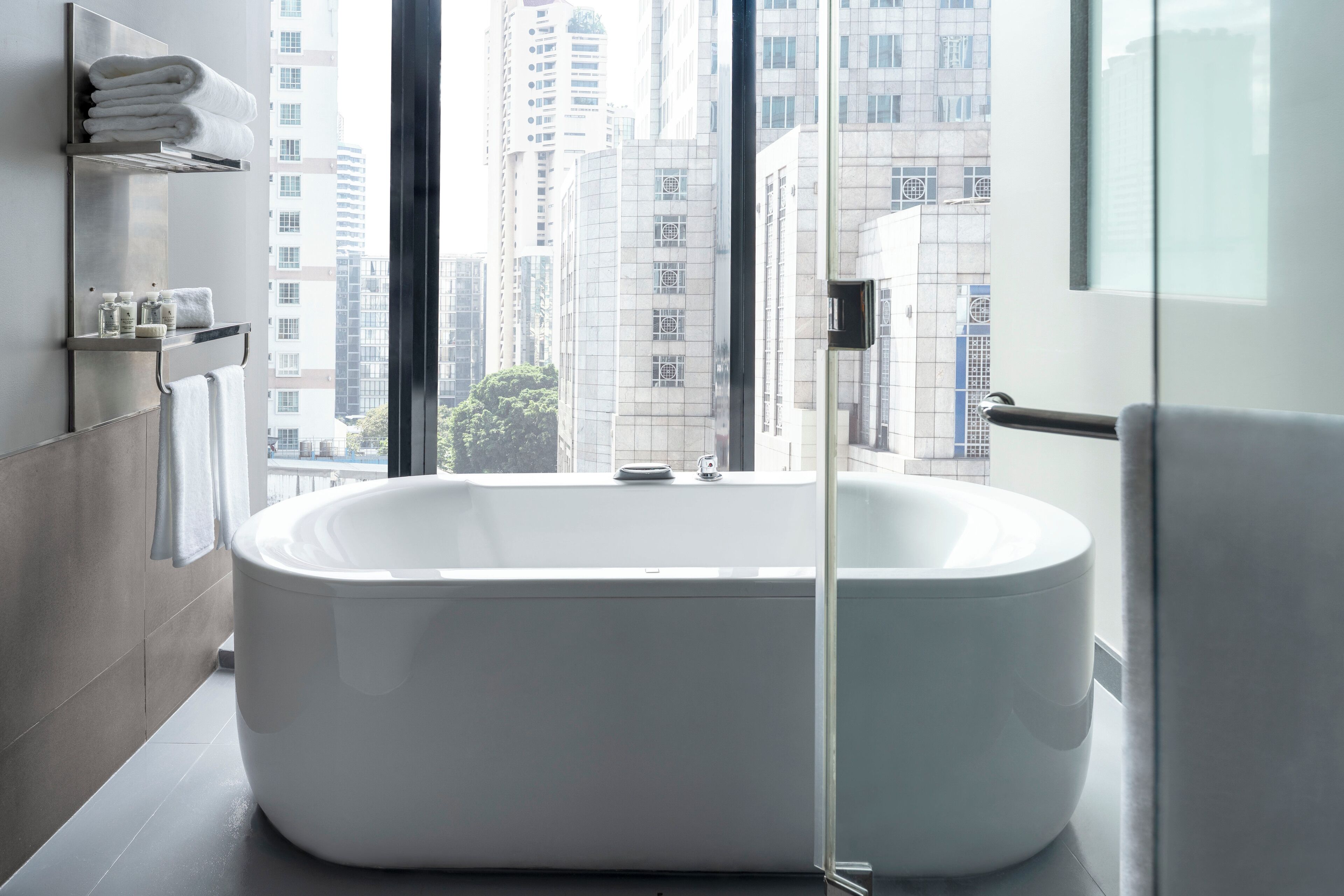 Un baño moderno con una bañera independiente, cabina de ducha de vidrio y vistas a altos edificios a través de amplios ventanales.