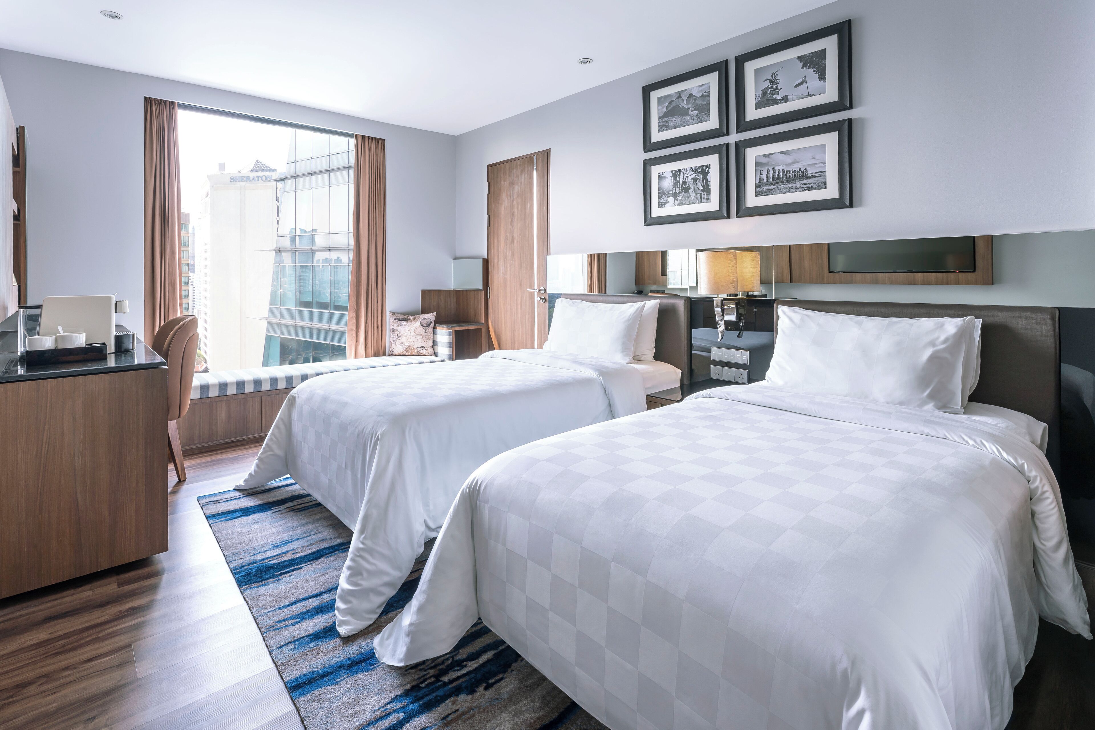 Une chambre d’hôtel avec deux lits, un bureau, une chaise, une lampe de bureau et une grande fenêtre avec des rideaux.