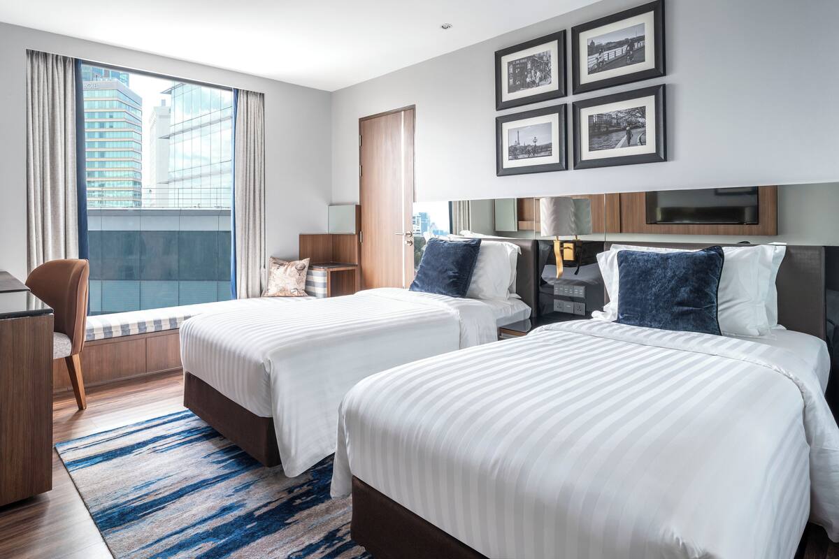 A hotel room with two beds, a desk, a chair, a large window with curtains, and framed pictures on the wall.