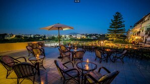 An outdoor terrace with round tables and chairs, a large umbrella, and a view of the cityscape at dusk. - Roxani Hotel (Malevizi)