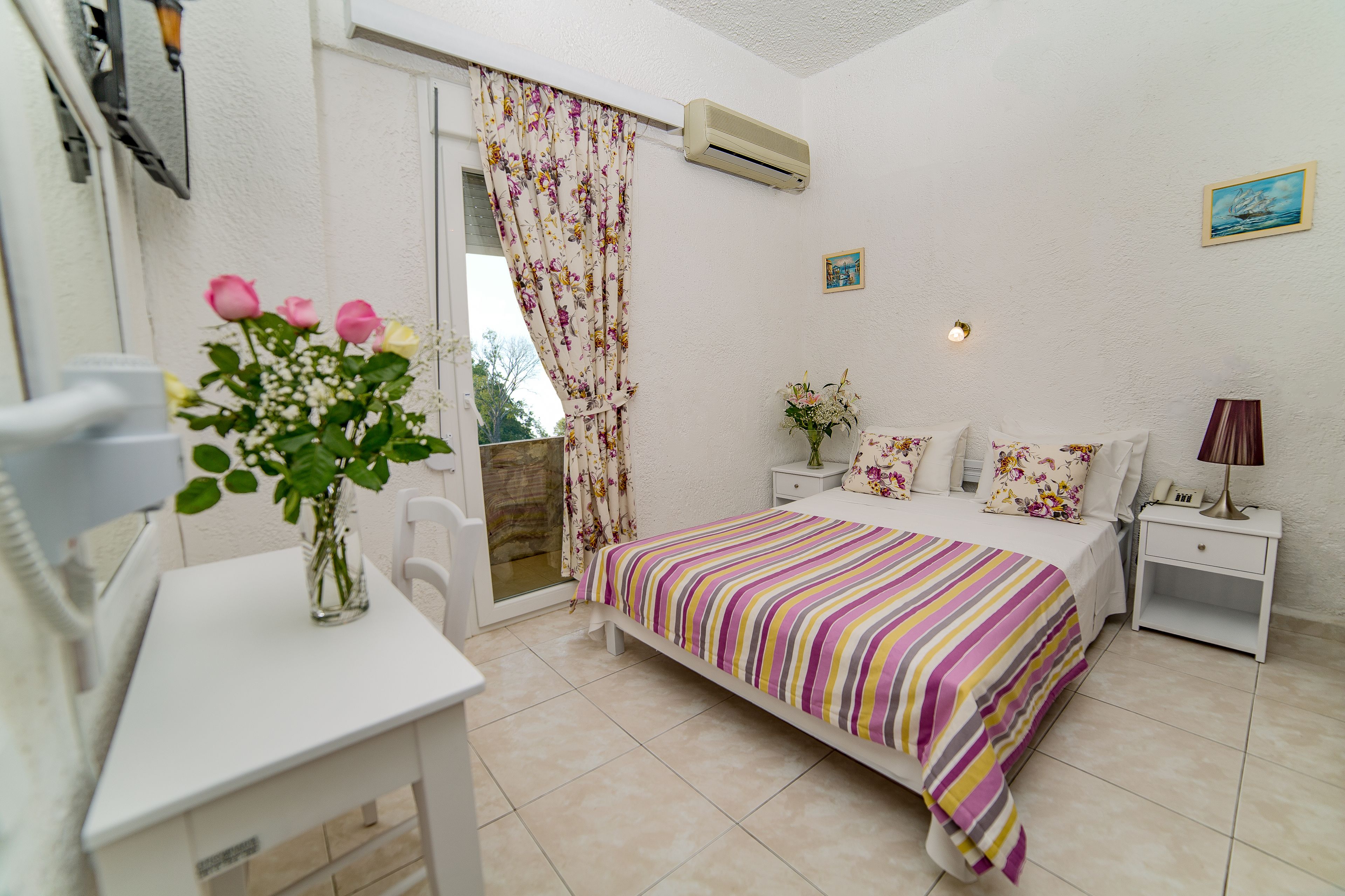 A bedroom with a bed, bedside tables, a desk, a chair, a window with curtains, and an air conditioning unit.