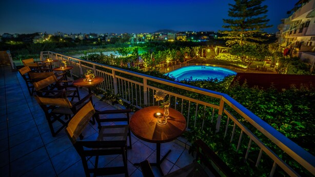 An outdoor terrace with tables and chairs, a pool, and a view of the city at night. - Roxani Hotel (Malevizi)