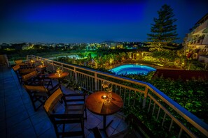 An outdoor terrace with tables and chairs, a pool, and a view of the city at night.