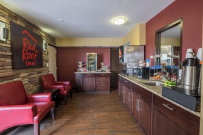 Un hall d’hôtel comprenant un comptoir d’accueil, un espace salon avec des fauteuils rouges, et un coin cuisine équipé d’un réfrigérateur et d’une machine à café.