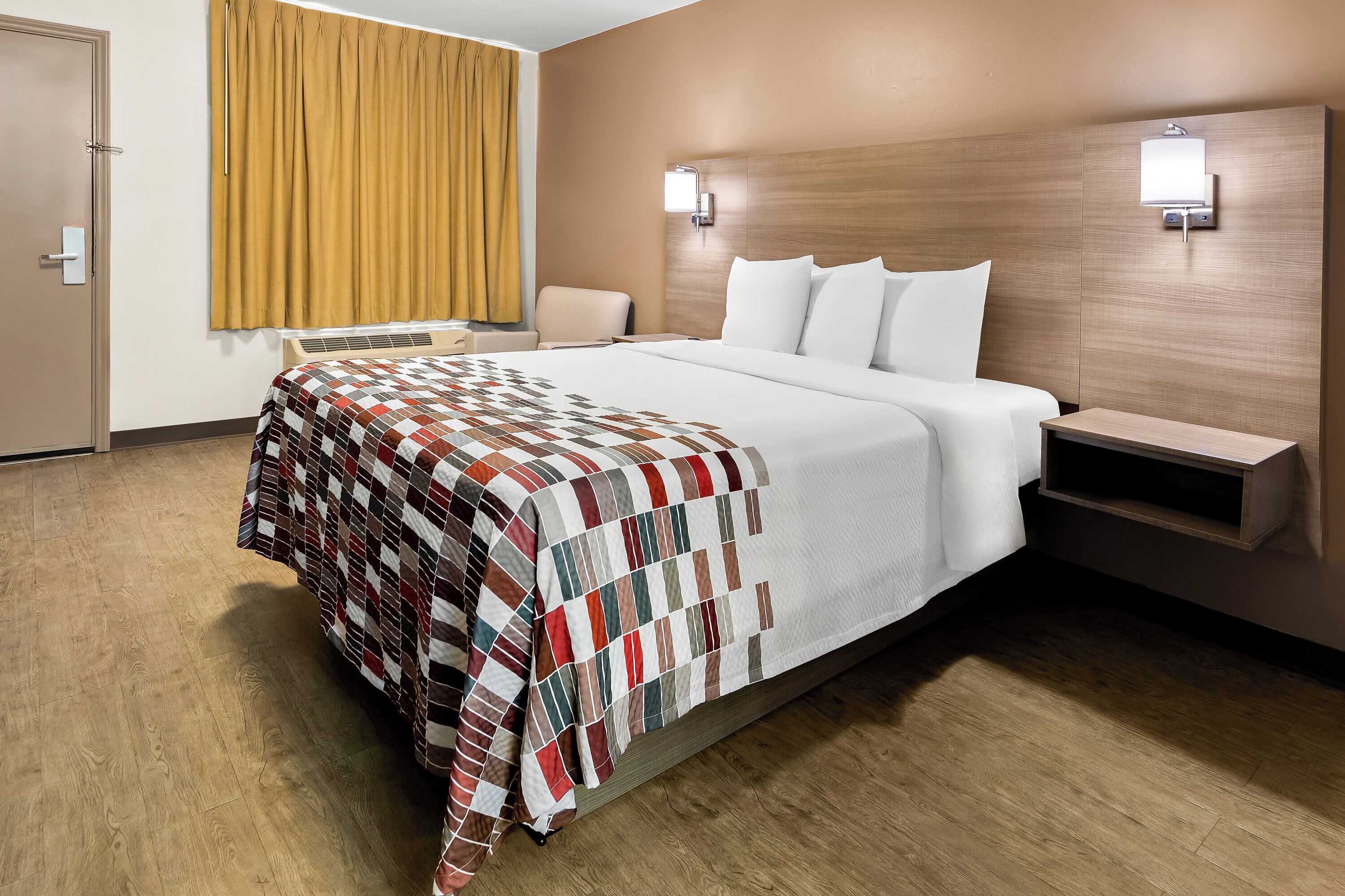 Hotelkamer met een groot bed, nachtkastje, stoel, raam met gordijnen en een wandlamp.
