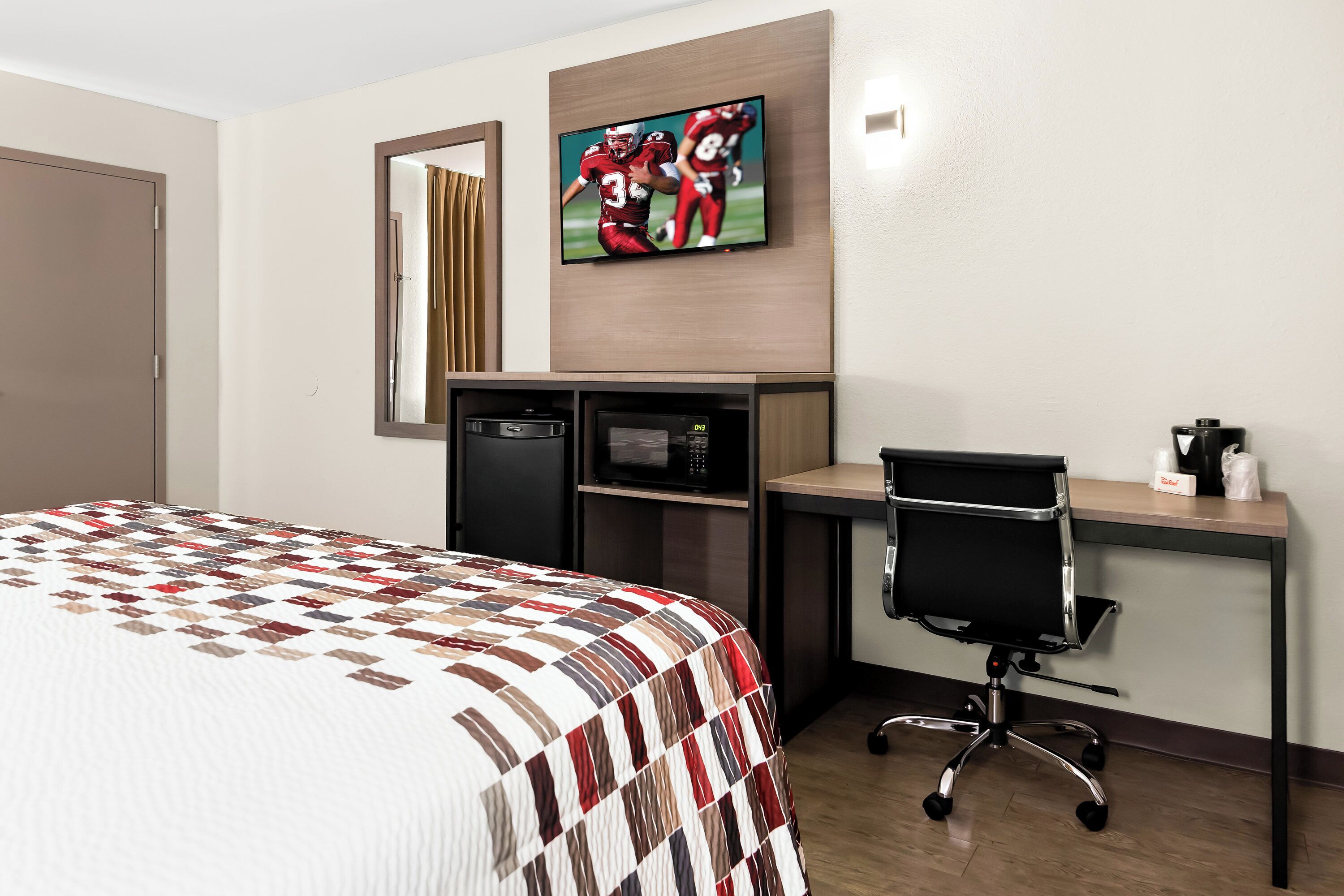Habitación de hotel con una cama, un escritorio con computadora, un microondas y un televisor mostrando un partido deportivo.
