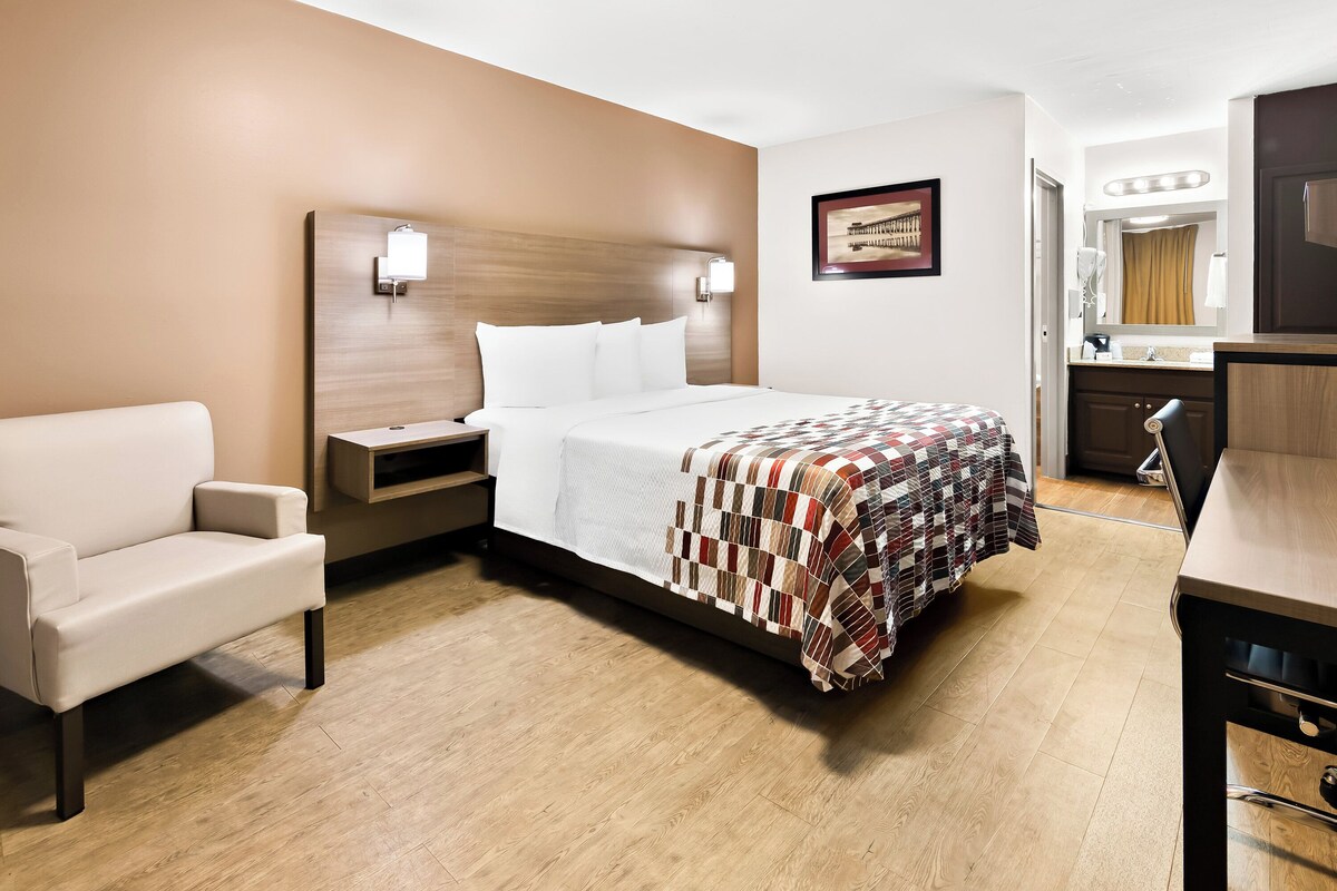 A hotel room with a bed, a chair, a desk, a nightstand, a mirror, and a bathroom with a sink and a shower.