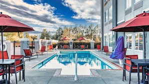 A swimming pool with red umbrellas and chairs, surrounded by a fence and trees.