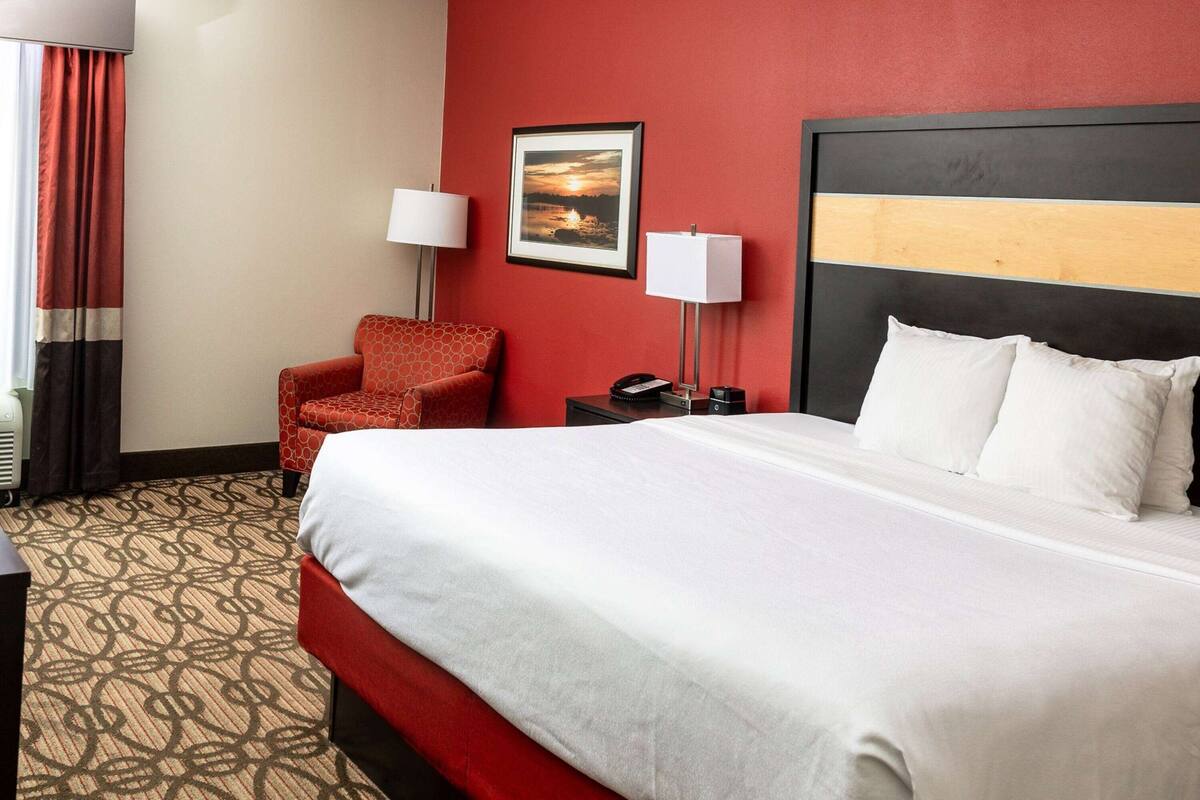 A hotel room with a large bed, a red armchair, a nightstand with a lamp, and a framed picture on the wall.