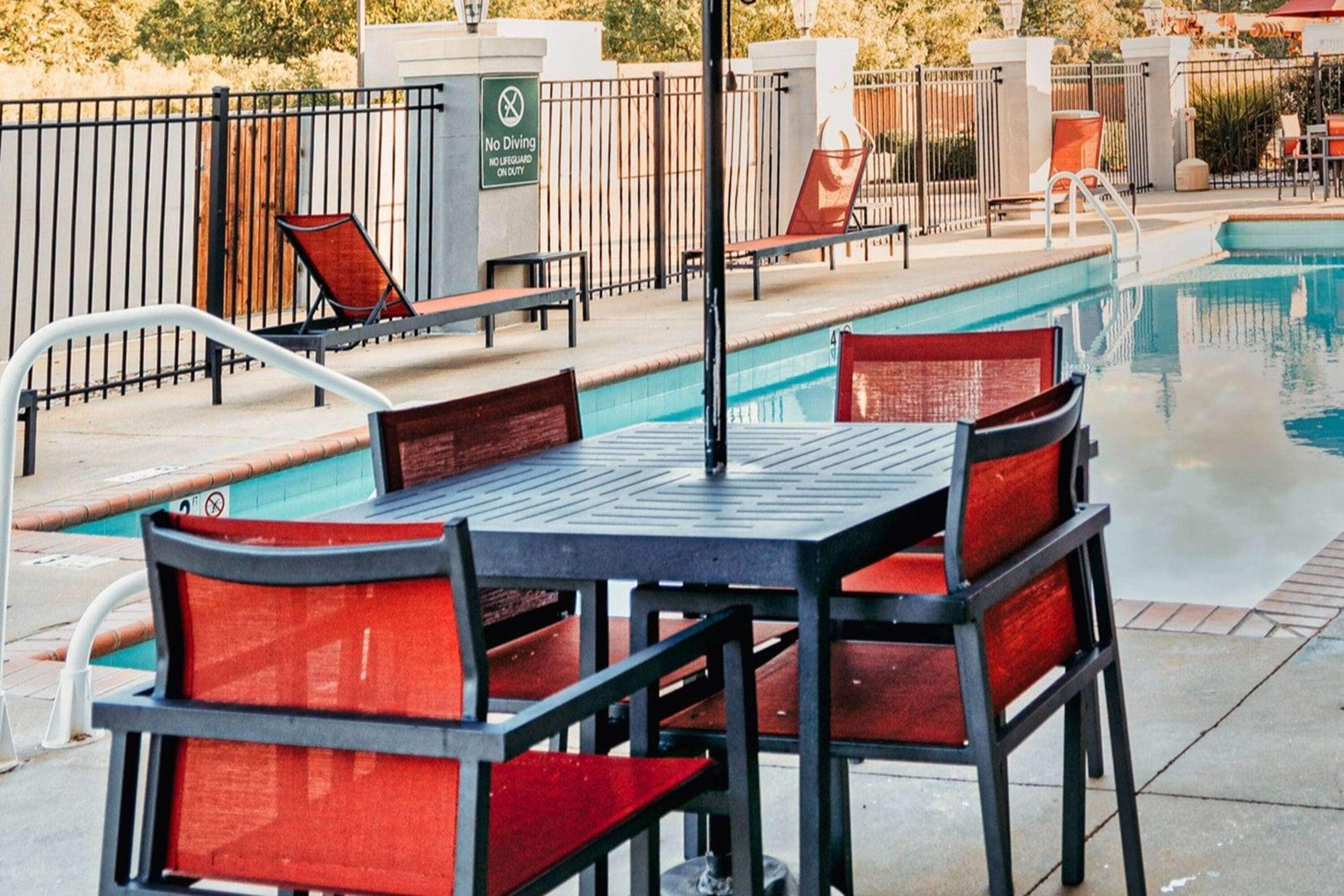 Zona junto a la piscina con mesa y sillas, un cartel que prohíbe los saltos y una piscina cercada.