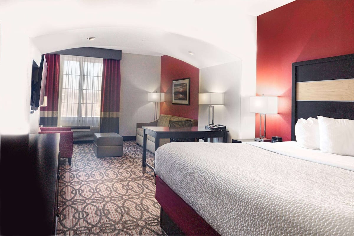 A hotel room with a large bed, a desk, a chair, a small table, a lamp, and a window with curtains.