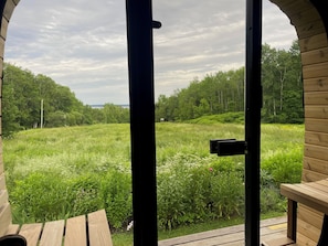 A wooden deck with a bench, a door leading to a lush green field, and a view of trees and a cloudy sky.