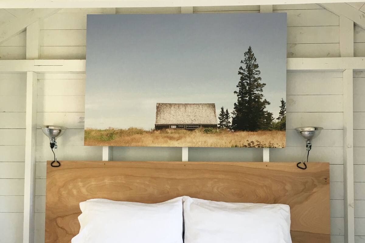 A bed with white bedding, a wooden headboard, and a painting of a barn and tree on the wall above.