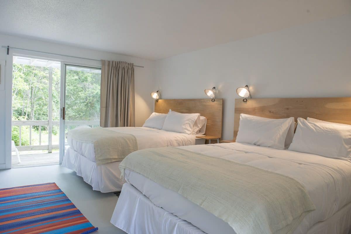 A hotel room with two beds, a large window with curtains, and a view of greenery outside.