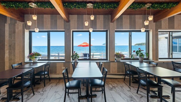 Ein Restaurant mit großen Fenstern, die Meerblick bieten, Holzbalken an der Decke und Topfpflanzen.