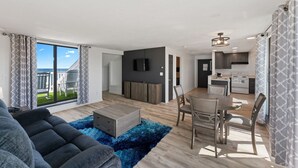 Ein modernes Wohnzimmer mit einer Couch, einem Couchtisch und einem Essbereich mit Tisch und Stühlen. Im Hintergrund ist eine Küche mit Schränken und einem Fernseher zu sehen.