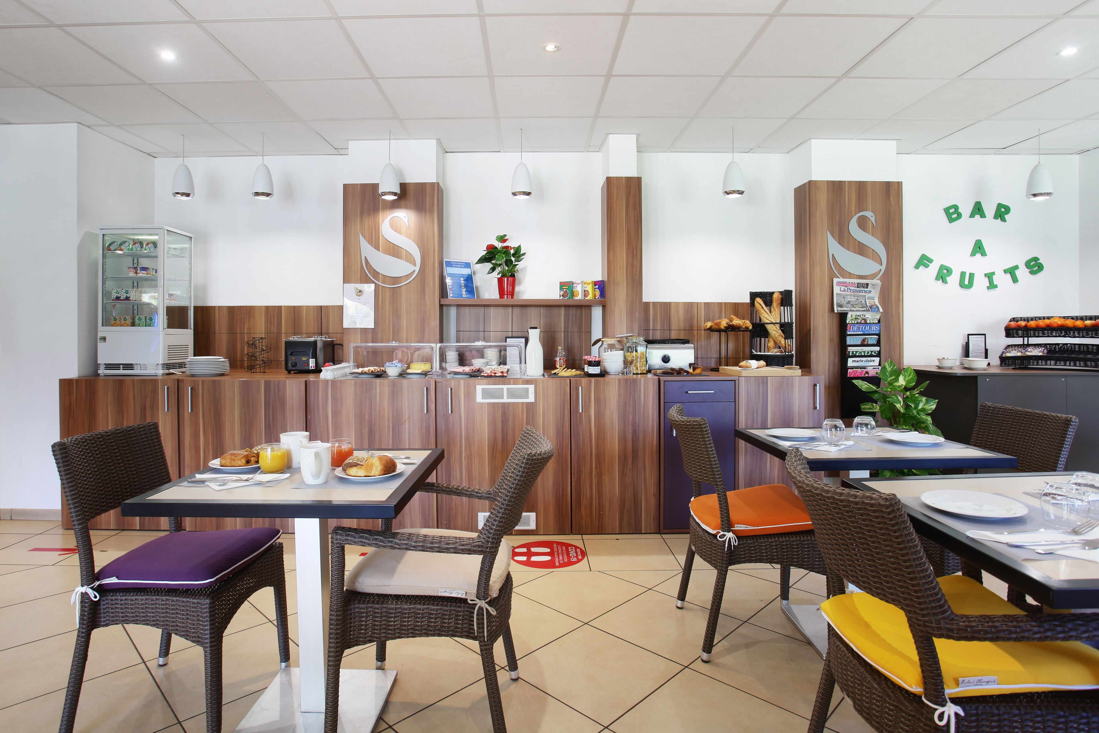 Un café moderno con detalles de madera y un letrero que dice 'Bar a Fruits', con mesas y sillas, una máquina de café y un refrigerador.