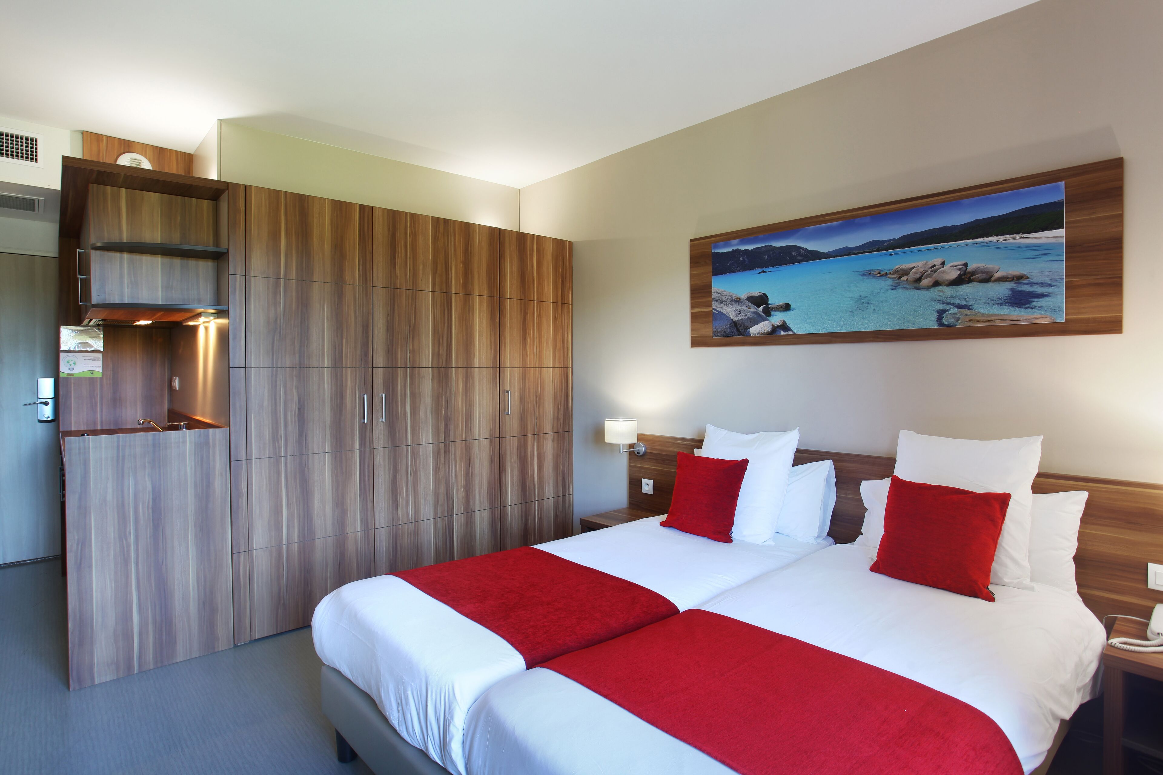 Habitación de hotel con una cama grande, un armario de madera, un cuadro de una playa y una mesita de noche con lámpara.