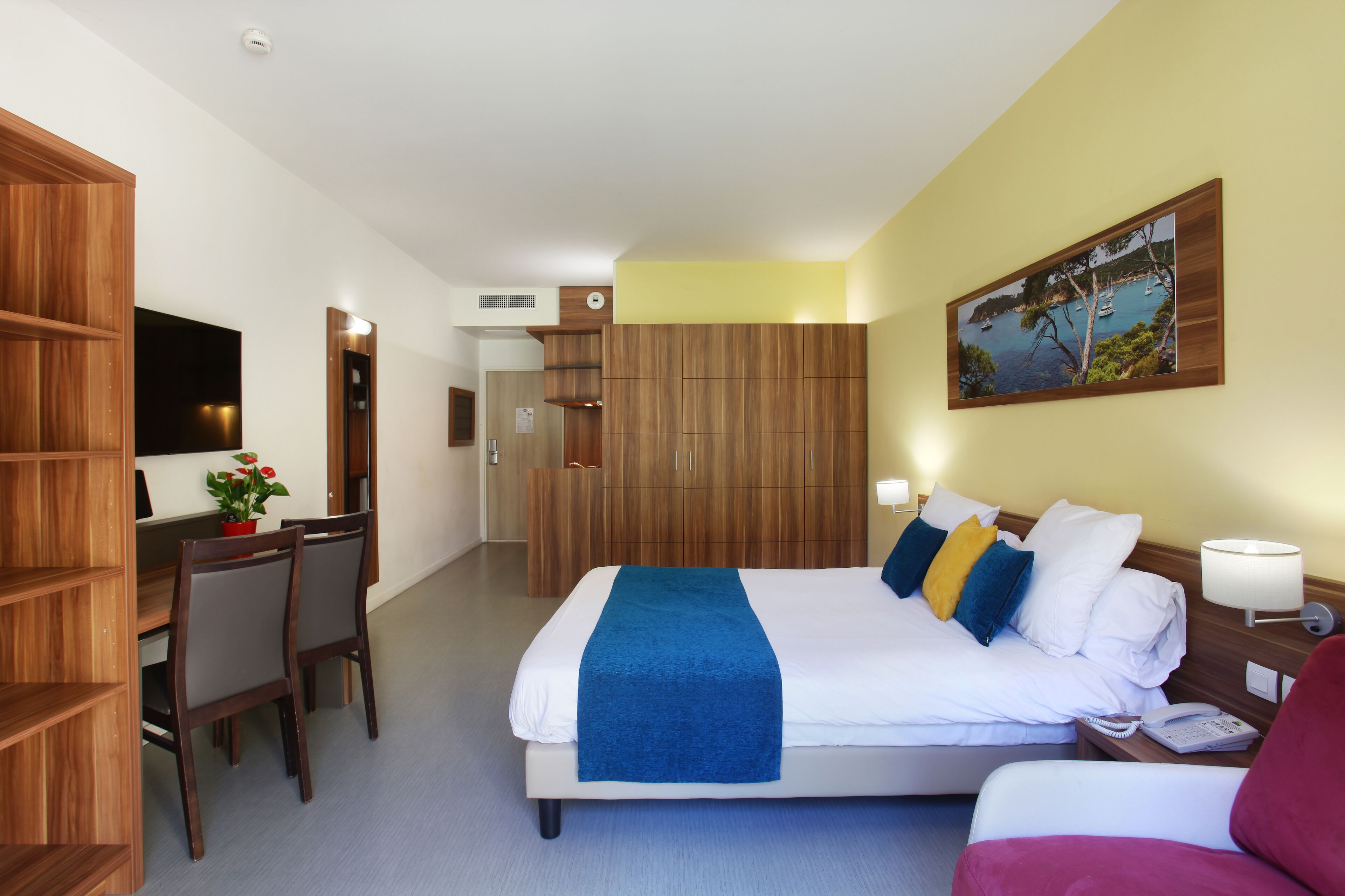 A hotel room with a bed, a desk, a chair, a bookshelf, and a painting on the wall.