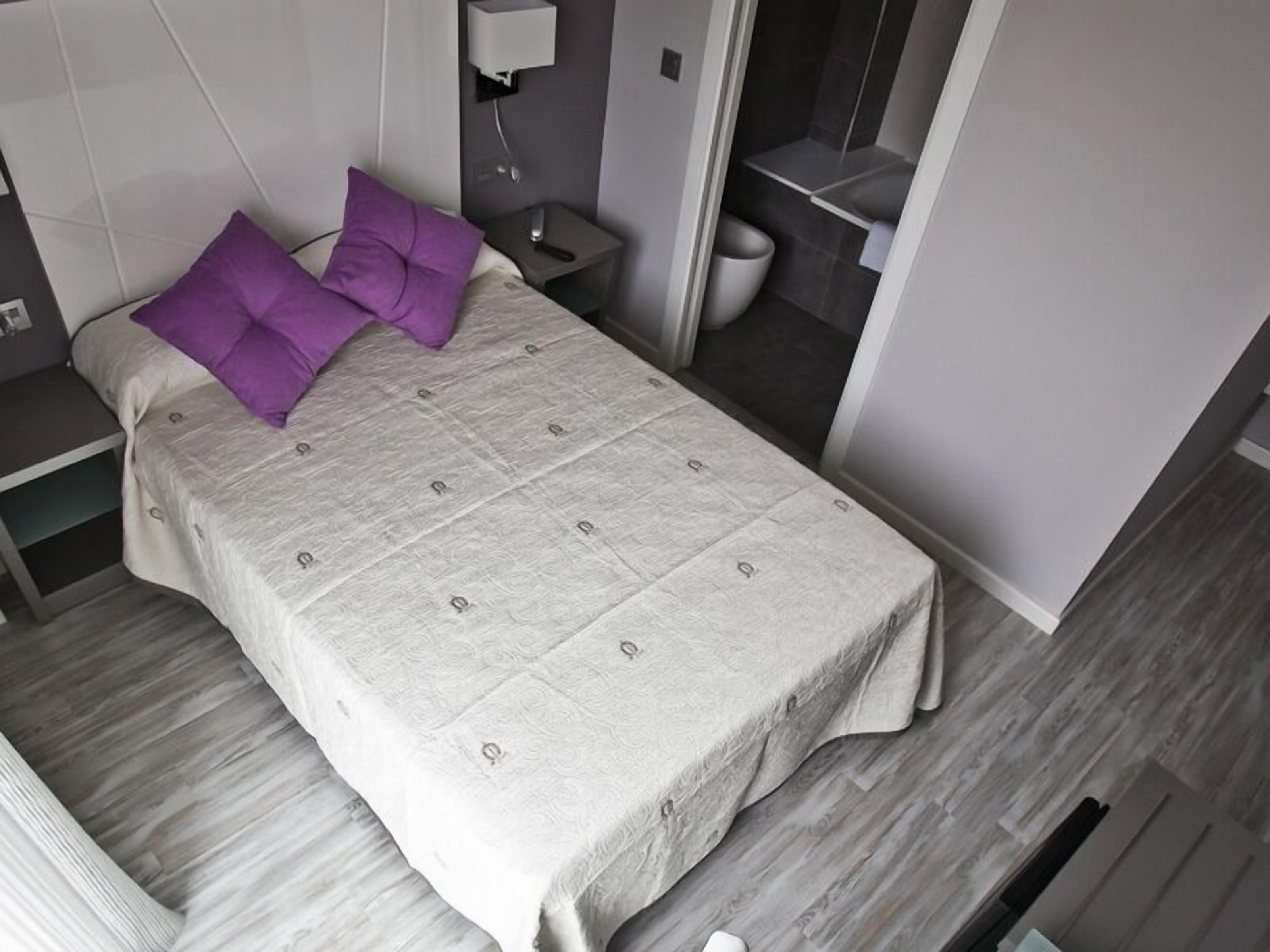 A modern bedroom with a bed, two purple pillows, a bedside table with a lamp, and a small bathroom visible through an open door.