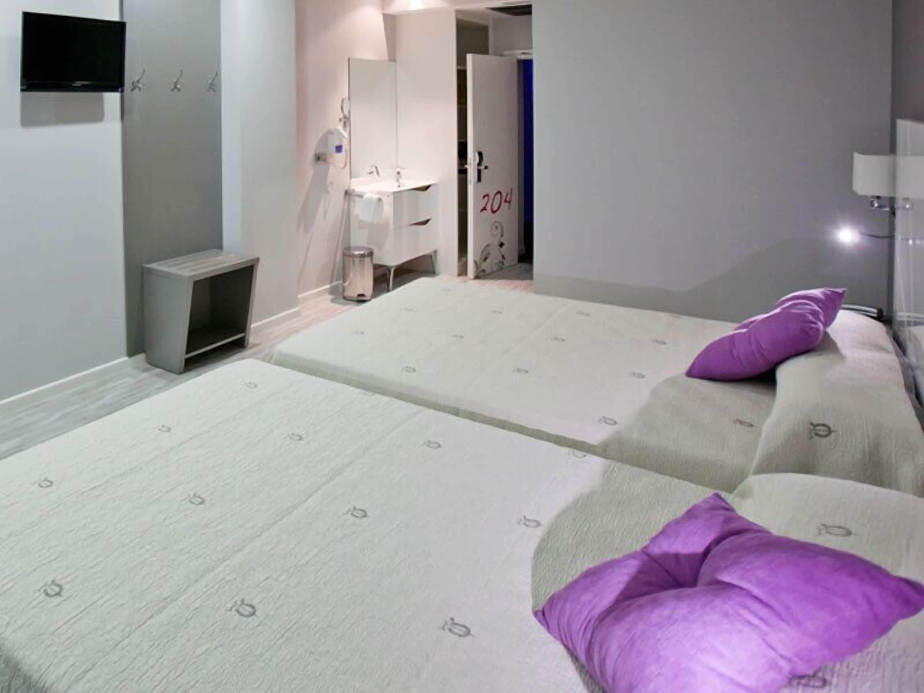 Habitación de hotel con dos camas, baño con espejo y lavamanos, y televisor montado en la pared.