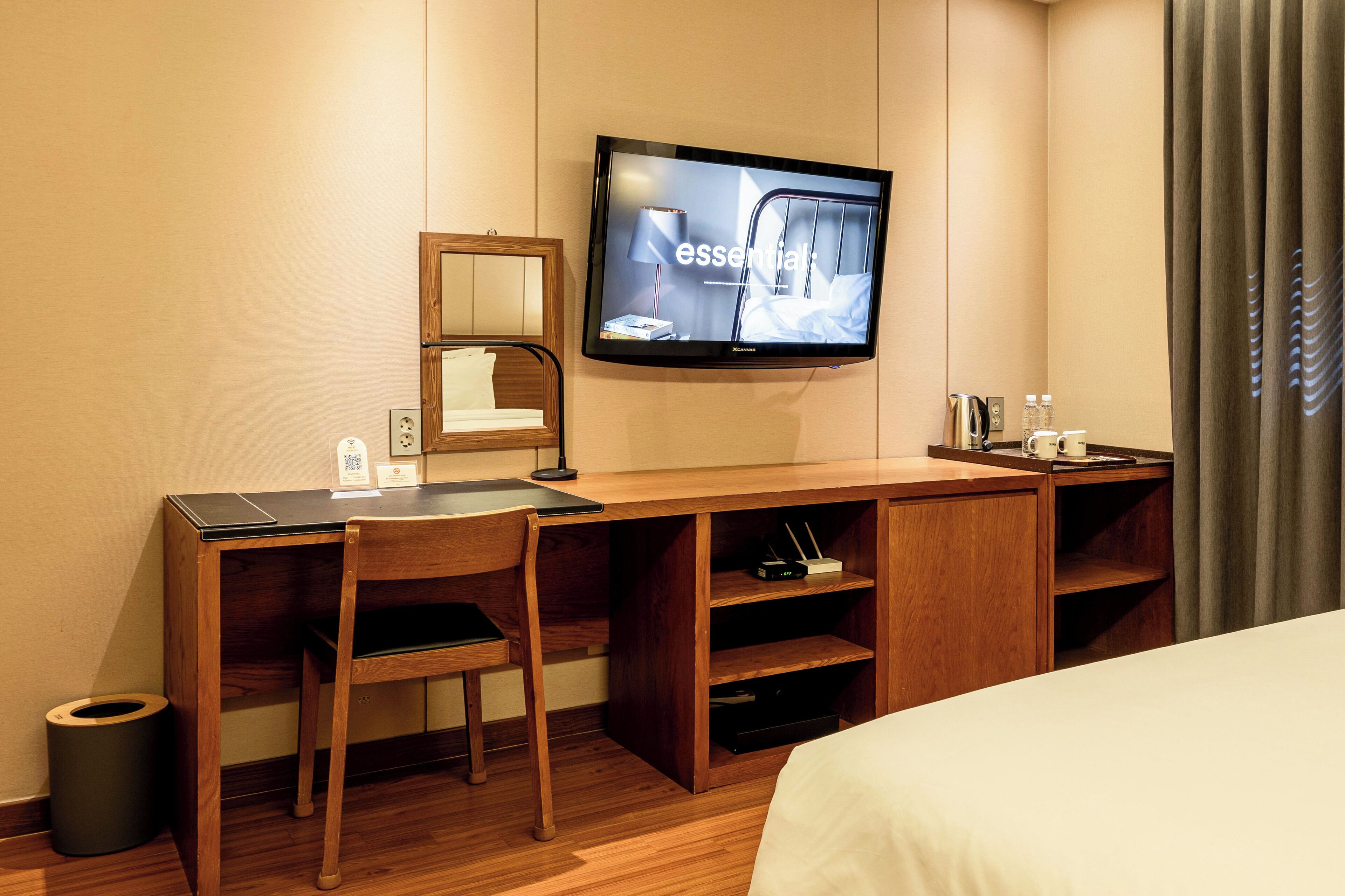 A hotel room with a wooden desk, a chair, a television mounted on the wall, a bed, a small shelf with toiletries, and a trash can.
