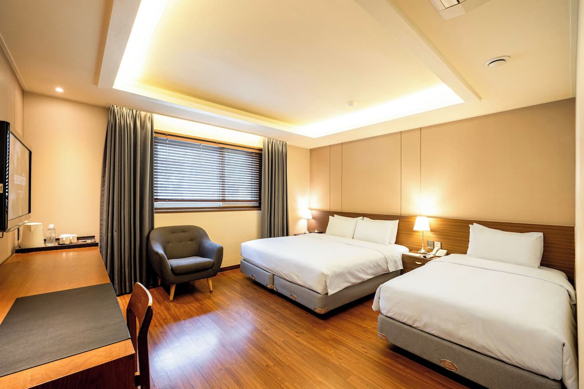 A hotel room with a desk, two beds, a chair, and a window with curtains.